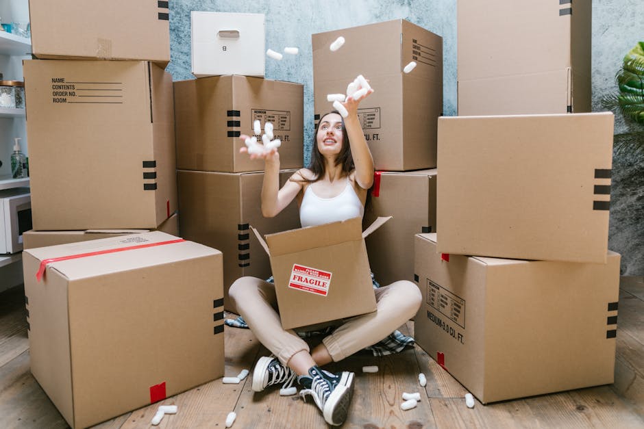 A young woman with light skin and long brown hair, sitting cross-legged on a wooden floor inside a home, surrounded by several large cardboard moving boxes, some labeled with fragile stickers. She is smiling and looking up as she tosses white packing foam into the air, with foam pieces scattered around her. The boxes vary in size, some stacked behind her and others placed on the floor, with additional packing materials and a plastic-wrapped item visible. The room has a textured blue wall and a green potted plant in the corner. This scene depicts the packing stage of house removals, where [COMPANY_NAME] assists in home relocation by managing packing and preparing items for transport, including careful handling of household furniture and fragile belongings during the loading process.
