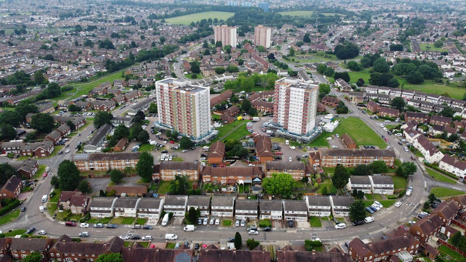Aerial view of a residential area with a mixture of high-rise apartment blocks and terraced houses in Southbury and Carterhatch estate, Enfield. The image shows a street with parked cars running along the front of the terraced houses, which have driveways and small front gardens. Behind these, there are several mid-sized apartment buildings with white facades and red accents, likely part of a home relocation or furniture transport project by Enfield Man and Van. Surrounding the area are green spaces with trees and grass, with additional houses and buildings extending towards the horizon. The scene is captured during daylight with overcast weather, offering a clear view of the neighborhood's layout, typical of local house removals and packing and moving processes.