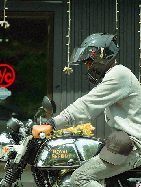 A person wearing a black full-face motorcycle helmet with a transparent visor, a light grey sweatshirt, and grey jeans is seated on a black Royal Enfield motorcycle, which is parked outside a building with vertical dark metal cladding. The individual’s right hand is gripping the handlebar, and they are wearing a black wristwatch. The motorcycle features a shiny chrome fuel tank with the Royal Enfield logo and is positioned on a paved surface. In the background, there is a partially visible door with a glass panel showing a blurred interior, and a red 'No Parking' sign is attached to the door frame. The scene appears to be related to a home relocation or moving process, with the motorcycle potentially used for transport or moving logistics, highlighting the connection to house removals and furniture transport services provided by Enfield Man and Van.