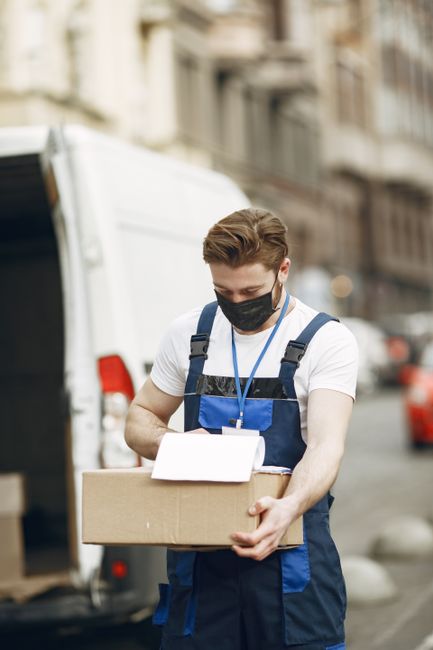 A young male mover, wearing a black face mask, a white T-shirt, and a dark blue uniform with reflective strips, is standing outdoors on a city street. He is holding a cardboard box with both hands, preparing to load or unload it as part of a home relocation or furniture transport process. Behind him, a white van is parked near a building with old architecture, and other vehicles are visible on the street. The scene is illuminated by natural daylight, with a slightly blurred background showing residential or commercial buildings. The image captures the professional packing and moving activities undertaken by Enfield Man and Van, supporting their house removals and relocation services in the Ponders End area.