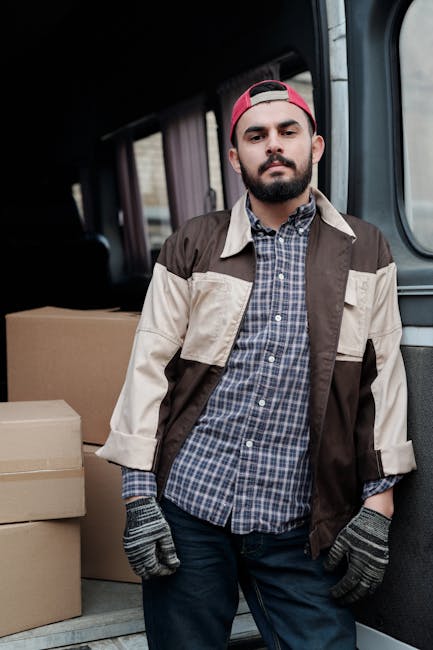 A young man with a beard and wearing a red cap, brown and beige work jacket, blue checkered shirt, and grey gloves, standing inside the back of a van used for home relocation. Behind him, several cardboard boxes are arranged on the van's floor, indicating loading or unloading activity for furniture transport and packing during a house move. The van interior is visible with dark lining and a partially open sliding door, suggesting an active loading process. Sunlight filters through the open vehicle, illuminating the scene clearly, which reflects the logistical aspects of professional removals offered by Enfield Man and Van in their house removals service for Ponders End high street moves, Enfield.