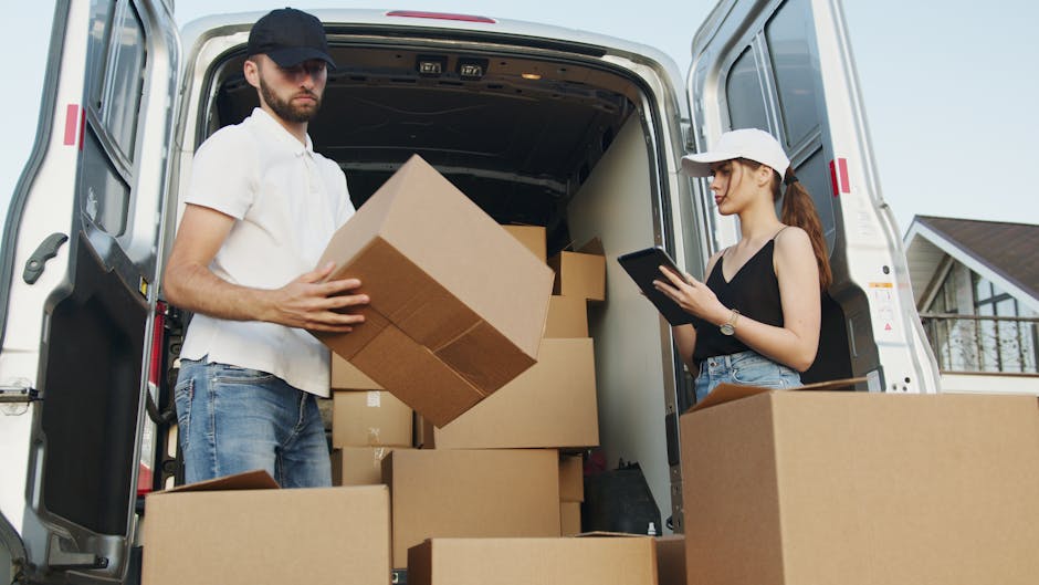 A young male mover, wearing a black face mask, a white T-shirt, and a dark blue uniform with reflective strips, is standing outdoors on a city street. He is holding a cardboard box with both hands, preparing to load or unload it as part of a home relocation or furniture transport process. Behind him, a white van is parked near a building with old architecture, and other vehicles are visible on the street. The scene is illuminated by natural daylight, with a slightly blurred background showing residential or commercial buildings. The image captures the professional packing and moving activities undertaken by Enfield Man and Van, supporting their house removals and relocation services in the Ponders End area.